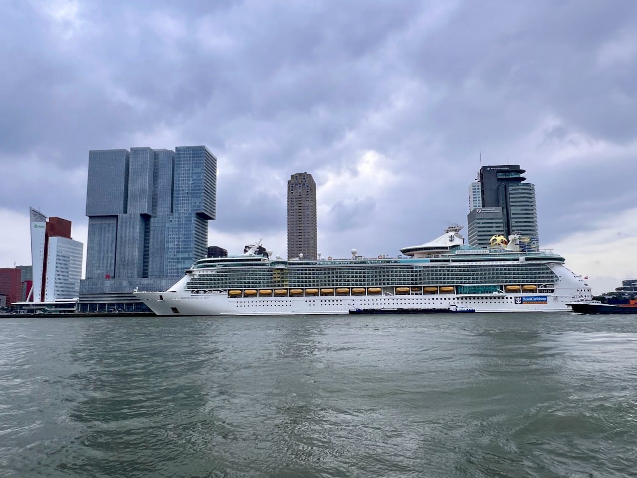 A cruise ship beside the modern skyline of Rotterdam, Netherlands, under cloudy skies.